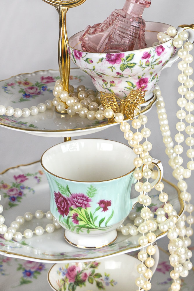 Vintage teacup with a vintage three tiered sweet/sandwich stand, antique perfume bottle, a vintage butterfly pin and a string of pearls.