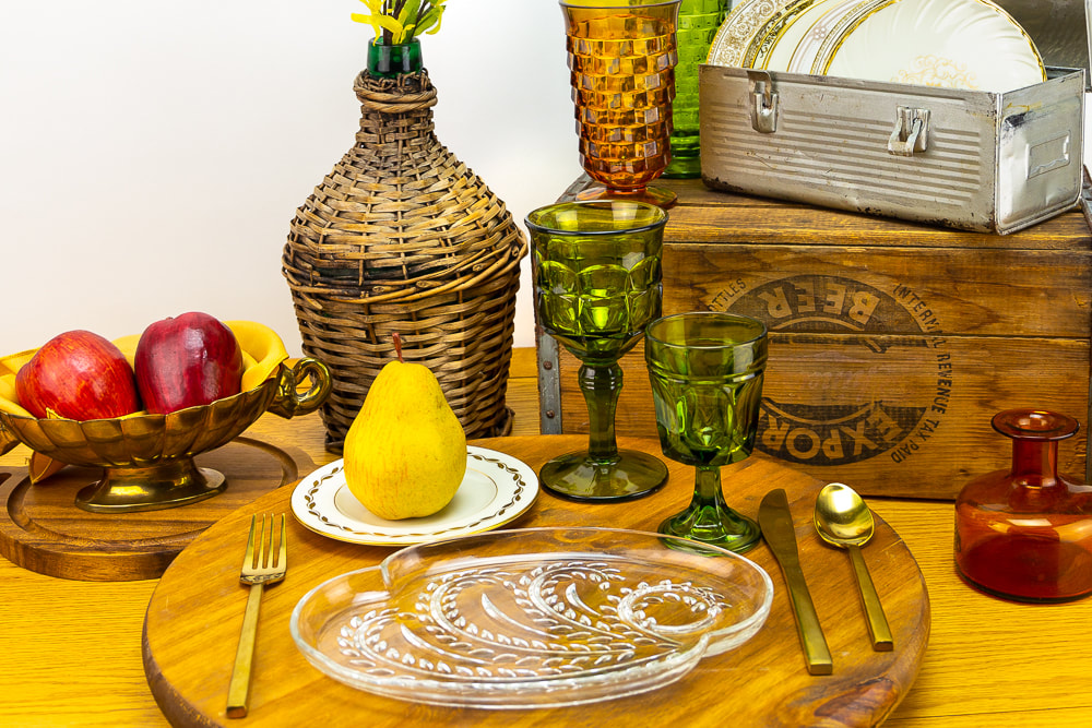 Vintage green goblet rentals mixed with other decor rentals of a wood crate, metal lunch box, gold flatware and a jug of wine covered with rattan.